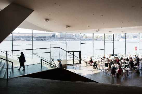 Binnenruimte van het Eye Filmmuseum met grote ramen, uitzicht op het water en mensen aan tafels.